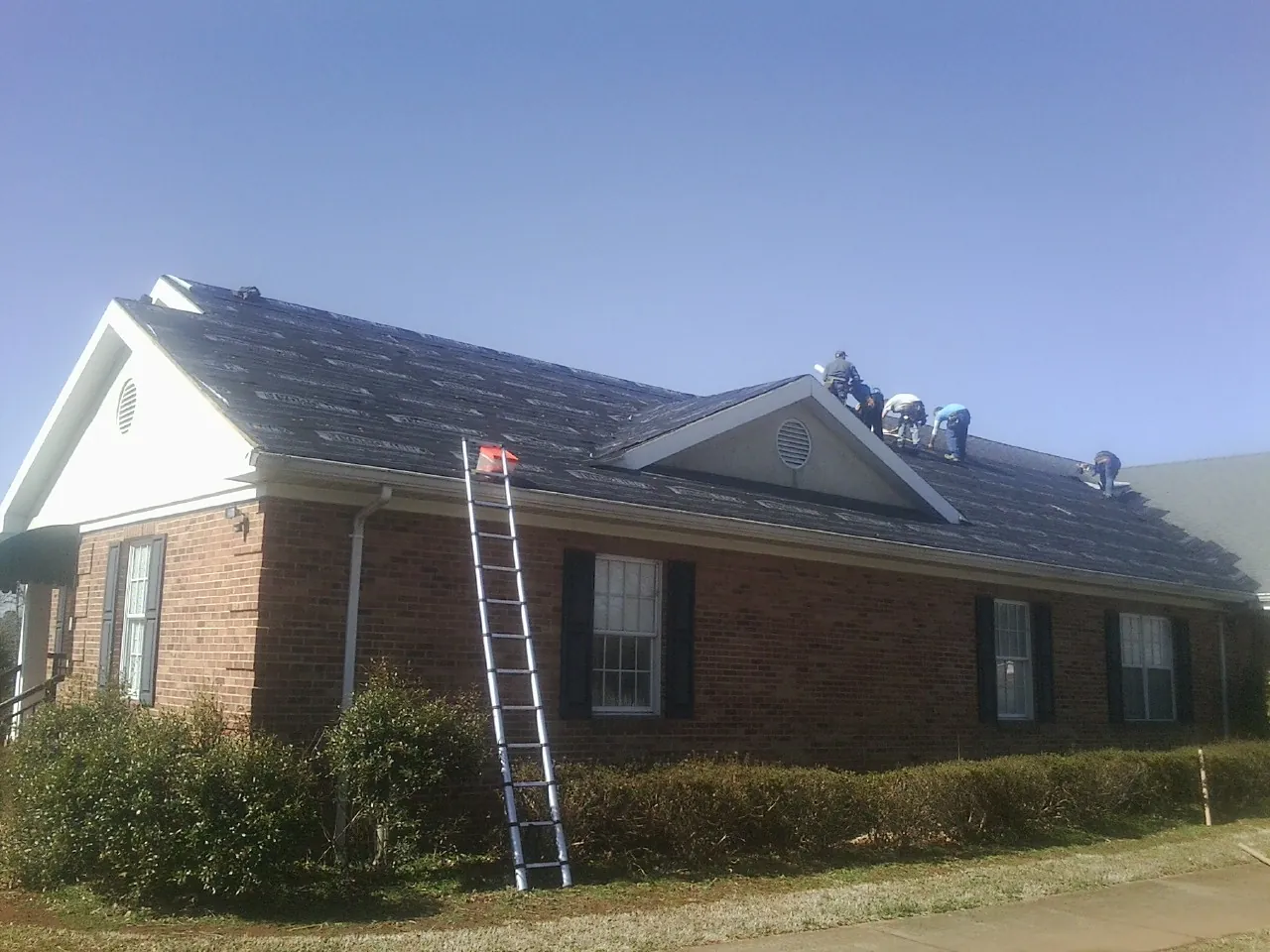 Crew tearing shingles off a brick ranch roof with a ladder set — Metal Roof Repair work by our crew in Grand Mound